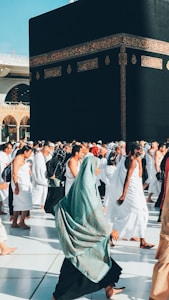 A large group of people walking around a black cuboid structure adorned with intricate gold patterns on its sides. The individuals are dressed in white robes, and a woman in a hijab is prominently walking in the foreground. The scene appears to be part of a significant cultural or religious event, taking place in a sunny, spacious plaza.