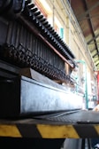 A close-up view of industrial machinery with pipes and metal components, positioned inside a large industrial space with high ceilings. The structure is complex and appears to be part of a larger mechanical assembly line.