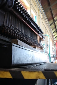 A close-up view of industrial machinery with pipes and metal components, positioned inside a large industrial space with high ceilings. The structure is complex and appears to be part of a larger mechanical assembly line.