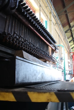 A close-up view of industrial machinery with pipes and metal components, positioned inside a large industrial space with high ceilings. The structure is complex and appears to be part of a larger mechanical assembly line.