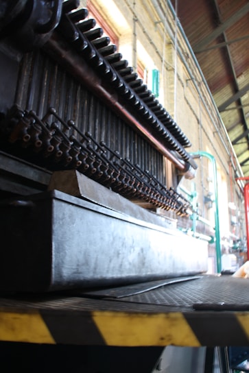 A close-up view of industrial machinery with pipes and metal components, positioned inside a large industrial space with high ceilings. The structure is complex and appears to be part of a larger mechanical assembly line.