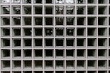 A grid of square glass blocks, each framed by white borders. The glass blocks are translucent with a textured surface, creating a pattern of reflections and refractions. The overall appearance is uniform and geometric, with a modern architectural style.