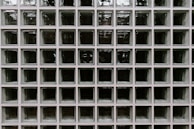 A grid of square glass blocks, each framed by white borders. The glass blocks are translucent with a textured surface, creating a pattern of reflections and refractions. The overall appearance is uniform and geometric, with a modern architectural style.