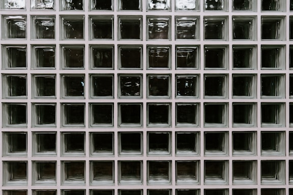 A grid of square glass blocks, each framed by white borders. The glass blocks are translucent with a textured surface, creating a pattern of reflections and refractions. The overall appearance is uniform and geometric, with a modern architectural style.