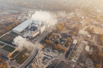 An aerial view of an industrial facility with multiple buildings and smoke emanating from chimneys. The area around the complex includes several construction sites, roads, and vehicles. The surrounding land has patches of vegetation, and the background fades into a townscape with scattered buildings and infrastructure.