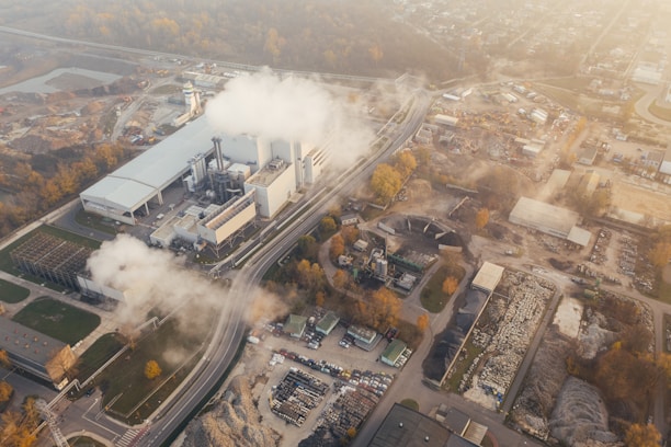 An aerial view of an industrial facility with multiple buildings and smoke emanating from chimneys. The area around the complex includes several construction sites, roads, and vehicles. The surrounding land has patches of vegetation, and the background fades into a townscape with scattered buildings and infrastructure.