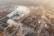 An aerial view of an industrial facility with multiple buildings and smoke emanating from chimneys. The area around the complex includes several construction sites, roads, and vehicles. The surrounding land has patches of vegetation, and the background fades into a townscape with scattered buildings and infrastructure.