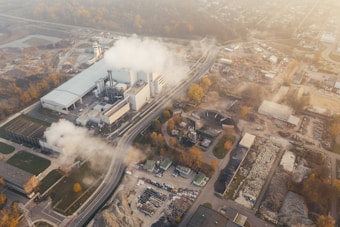 An aerial view of an industrial facility with multiple buildings and smoke emanating from chimneys. The area around the complex includes several construction sites, roads, and vehicles. The surrounding land has patches of vegetation, and the background fades into a townscape with scattered buildings and infrastructure.