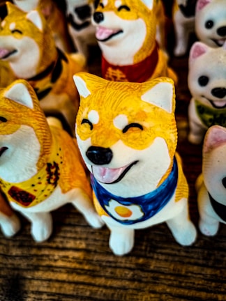 A collection of Shiba Inu figurines, with detailed features like smiling faces and decorative bandanas. The central figure has a blue bandana with an egg design, while others have different colored bandanas. The overall atmosphere is playful and whimsical.