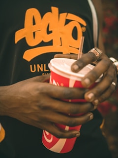 A person holding a branded red soda cup with a straw. The person is wearing a black shirt with a bold orange logo that reads 'Ecko Unlimited'. Several rings are visible on the person's fingers.