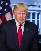 A middle-aged man with blonde hair is wearing a dark suit and a red tie. He is standing in front of a backdrop featuring the American flag and a building with columns, suggesting a governmental setting.