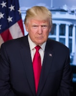 A middle-aged man with blonde hair is wearing a dark suit and a red tie. He is standing in front of a backdrop featuring the American flag and a building with columns, suggesting a governmental setting.
