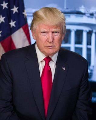 A middle-aged man with blonde hair is wearing a dark suit and a red tie. He is standing in front of a backdrop featuring the American flag and a building with columns, suggesting a governmental setting.