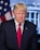 A middle-aged man with blonde hair is wearing a dark suit and a red tie. He is standing in front of a backdrop featuring the American flag and a building with columns, suggesting a governmental setting.