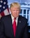 A middle-aged man with blonde hair is wearing a dark suit and a red tie. He is standing in front of a backdrop featuring the American flag and a building with columns, suggesting a governmental setting.