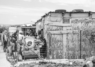 A group of people gather in front of a weathered wooden structure with peeling paint. The structure has a corrugated metal roof with car tires on top. There is a wire fence in the foreground, and various items are piled up against it, including a stroller. The scene portrays an informal settlement area.