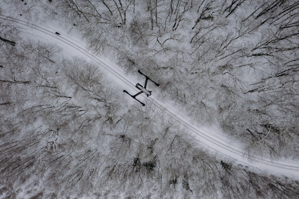 Aerial view of a snow-covered forest with bare trees. A small, dark drone is flying along a winding path that cuts through the trees. The scene is serene, with the snow blanketing the ground and branches.
