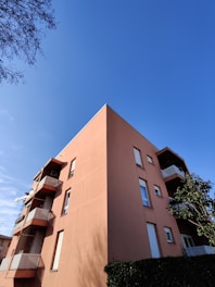 A multistory residential building with a modern design, featuring a salmon-colored exterior with balconies and rectangular windows against a clear blue sky. The building is partially obscured by a lush green hedge and a small tree on the right side.