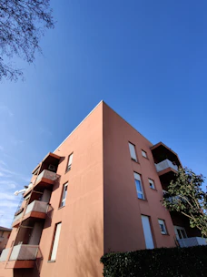 A multistory residential building with a modern design, featuring a salmon-colored exterior with balconies and rectangular windows against a clear blue sky. The building is partially obscured by a lush green hedge and a small tree on the right side.