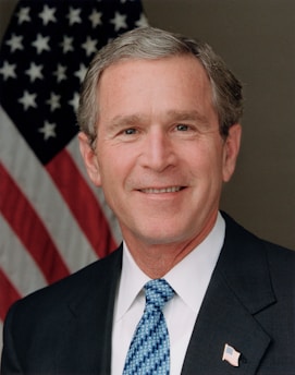 A man in a suit and tie stands in front of an American flag. He has short gray hair and is smiling.