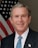 A man in a suit and tie stands in front of an American flag. He has short gray hair and is smiling.