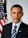 A man in a suit and tie is standing in front of an American flag and a blue curtain with a government seal partially visible. He has a serious and composed expression on his face.