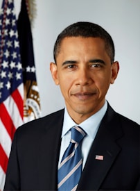 A man in a suit and tie is standing in front of an American flag and a blue curtain with a government seal partially visible. He has a serious and composed expression on his face.