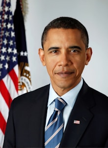 A man in a suit and tie is standing in front of an American flag and a blue curtain with a government seal partially visible. He has a serious and composed expression on his face.
