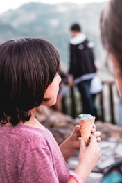 A child with short dark hair holds an ice cream cone with blue ice cream while standing outdoors. The child is wearing a pink shirt and a pink bracelet, gazing towards a blurred background that includes other people and a landscape.