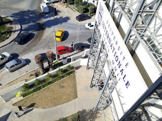 A large signboard with French text is situated on the side of a multi-story building. Several vehicles, including a yellow taxi and a red car, are stopped at an intersection. The area is landscaped with grass and small trees, and there are palm trees in the foreground. Pedestrians are visible walking on the sidewalks.