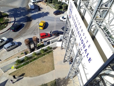 A large signboard with French text is situated on the side of a multi-story building. Several vehicles, including a yellow taxi and a red car, are stopped at an intersection. The area is landscaped with grass and small trees, and there are palm trees in the foreground. Pedestrians are visible walking on the sidewalks.