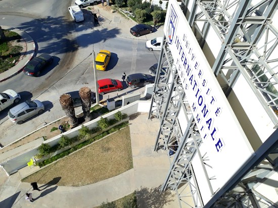 A large signboard with French text is situated on the side of a multi-story building. Several vehicles, including a yellow taxi and a red car, are stopped at an intersection. The area is landscaped with grass and small trees, and there are palm trees in the foreground. Pedestrians are visible walking on the sidewalks.