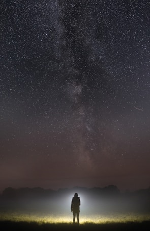 A person stands in an open field at night, illuminated by a beam of light from a flashlight. Above, the sky is filled with stars, showcasing the Milky Way galaxy. The scene evokes a sense of wonder and exploration, set against a backdrop of vast darkness.