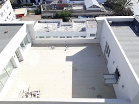 A spacious rooftop area with a tiled surface is surrounded by white walls and windows. In the background, several buildings and a palm tree are visible. Near the edge of the rooftop, some construction materials are scattered, and a small section of the rooftop is shaded.