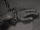A human hand is bound by thick, heavy chains, which are fastened with a lock. The lighting and shadows cast a gloomy and somber atmosphere.