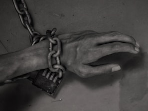 A human hand is bound by thick, heavy chains, which are fastened with a lock. The lighting and shadows cast a gloomy and somber atmosphere.