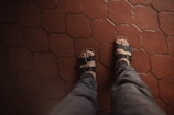 A pair of feet wearing black sandals is seen from above on a patterned red-tiled floor. The sandals have adjustable straps and reveal the toes. The pants worn are gray, suggesting a casual or relaxed setting.