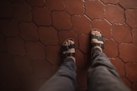 A pair of feet wearing black sandals is seen from above on a patterned red-tiled floor. The sandals have adjustable straps and reveal the toes. The pants worn are gray, suggesting a casual or relaxed setting.