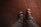 A pair of feet wearing black sandals is seen from above on a patterned red-tiled floor. The sandals have adjustable straps and reveal the toes. The pants worn are gray, suggesting a casual or relaxed setting.