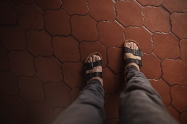 A pair of feet wearing black sandals is seen from above on a patterned red-tiled floor. The sandals have adjustable straps and reveal the toes. The pants worn are gray, suggesting a casual or relaxed setting.