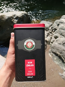 A hand is holding a dark cylindrical container with a red lid. The container has a label featuring a circular logo and reads 'Sıcak Çikolata' which means hot chocolate. The background shows a rocky riverbank with flowing water reflecting sunlight.