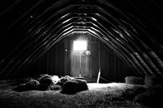 An attic with a high, arched ceiling constructed of wooden beams. Hay or straw bales are scattered on the floor, and light filters through a small window, casting shadows throughout the room. The atmosphere is rustic and slightly eerie.