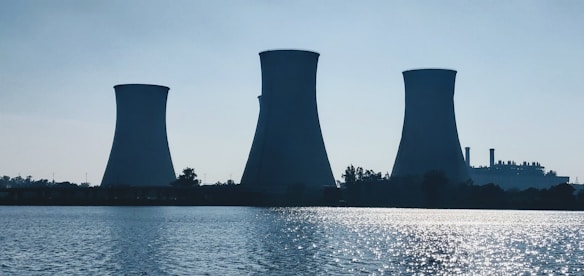 Three large cooling towers of a power plant stand near a body of water with trees surrounding them. The sky appears clear, and the calm water reflects sunlight.