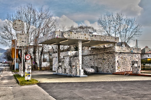 A gas station entirely covered with newspaper clippings, including the pumps and the building itself. The scene is set on a quiet street with a few bare trees and nearby houses. The sky is overcast, creating a somewhat muted atmosphere.