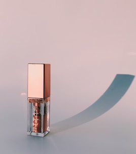 A stylish lip gloss container with a metallic pink cap and transparent body stands on a smooth surface casting a soft, elongated shadow.
