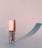 A stylish lip gloss container with a metallic pink cap and transparent body stands on a smooth surface casting a soft, elongated shadow.