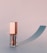 A stylish lip gloss container with a metallic pink cap and transparent body stands on a smooth surface casting a soft, elongated shadow.