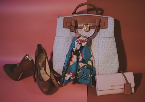 A collection of stylish accessories including a large handbag with a branded pattern, a pair of high-heeled shoes in a dark brown color, a floral scarf with vibrant colors, and a matching light pink clutch wallet. The background features a gradient of warm tones creating a fashionable and elegant display.