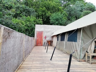 A wooden walkway leads to a canvas tent set up against a lush, green forest backdrop. The tent has several windows and is connected to a woven bamboo wall on one side. A wooden door and a fire extinguisher are visible at the end of the walkway, adding elements of utility and safety.