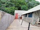 A wooden walkway leads to a canvas tent set up against a lush, green forest backdrop. The tent has several windows and is connected to a woven bamboo wall on one side. A wooden door and a fire extinguisher are visible at the end of the walkway, adding elements of utility and safety.